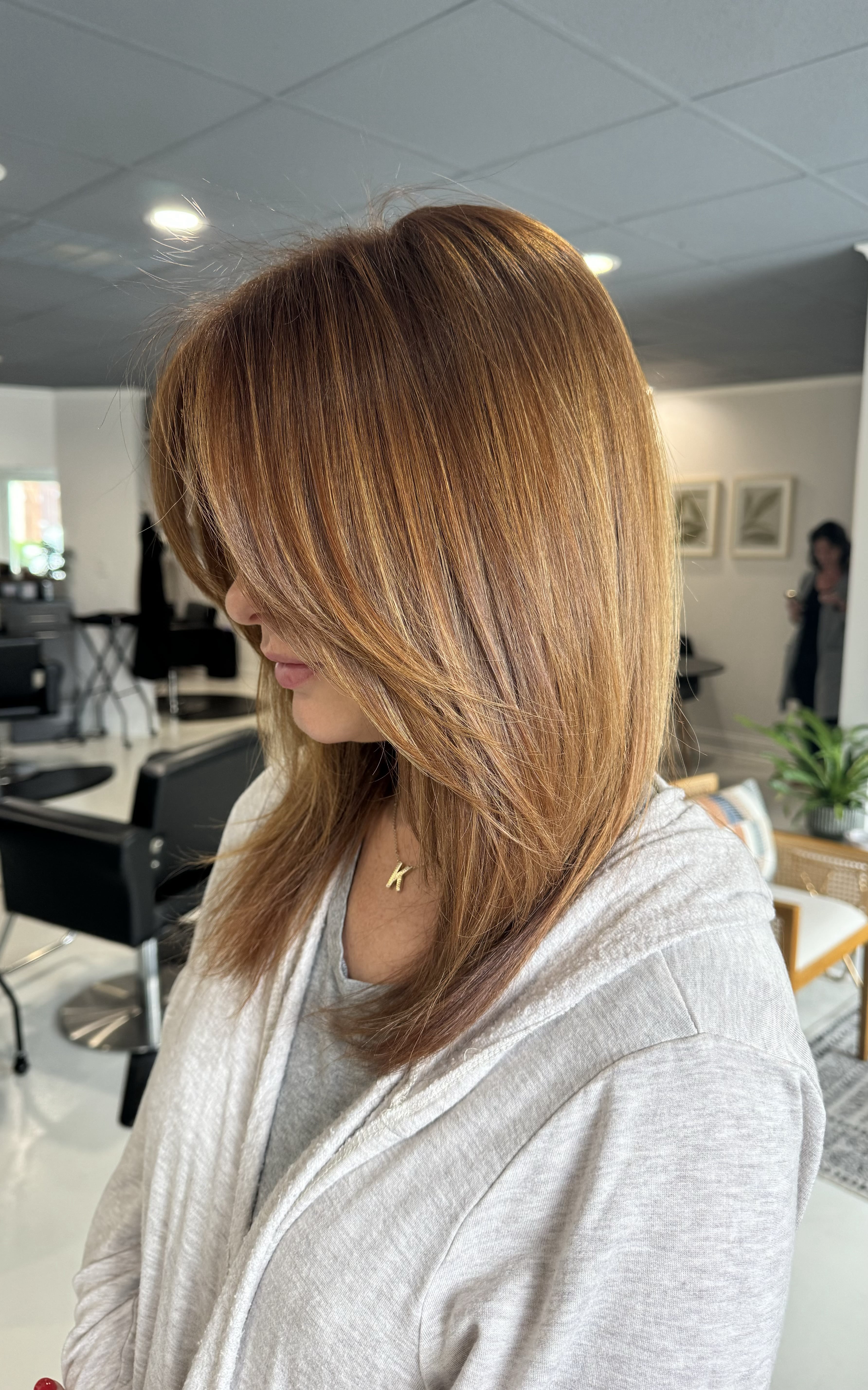 Side profile of a woman with medium-length, layered honey-blonde hair in a brightly lit salon setting