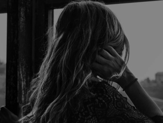 lack and white back view of a person with long wavy hair looking out a window with their hand to their head